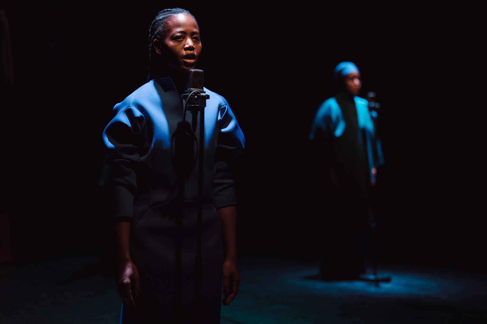 Two people stand on a dark stage and sing into microphones.