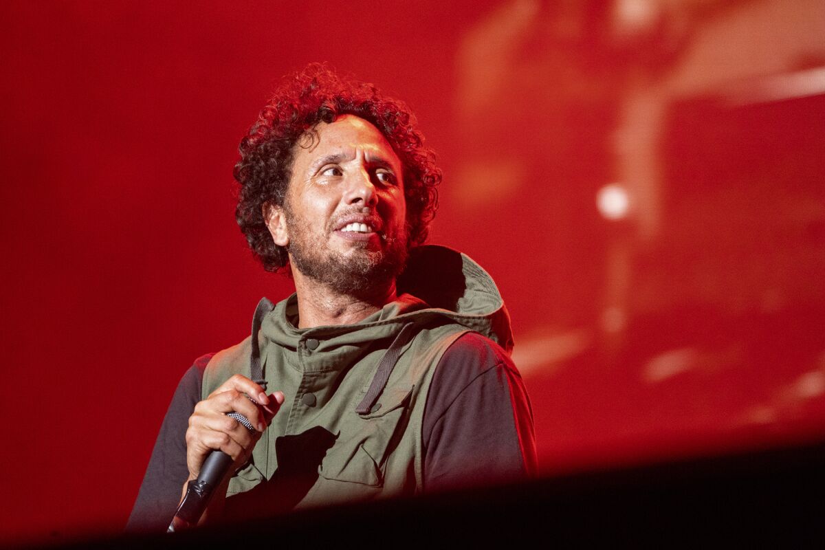 A man with curly hair holding a microphone on a stage against a red background