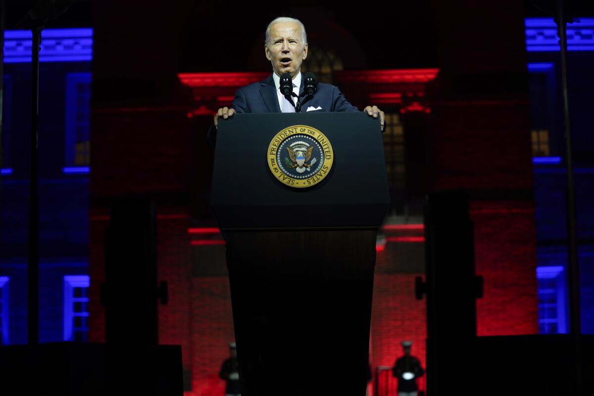 A man in a suit speaks at a lectern with the presidential seal, with illuminated buildings behind him