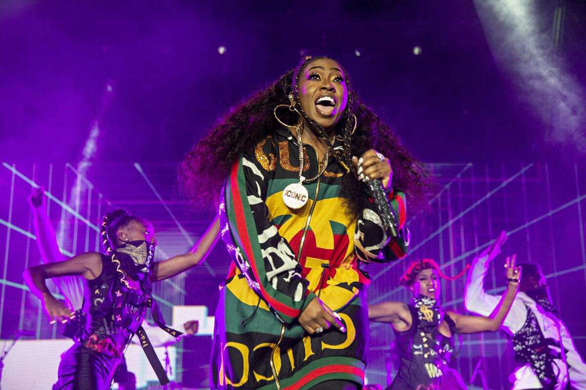 Missy Elliott in a yellow, green and red jumpsuit onstage holding a microphone. People in black outfits dance behind her