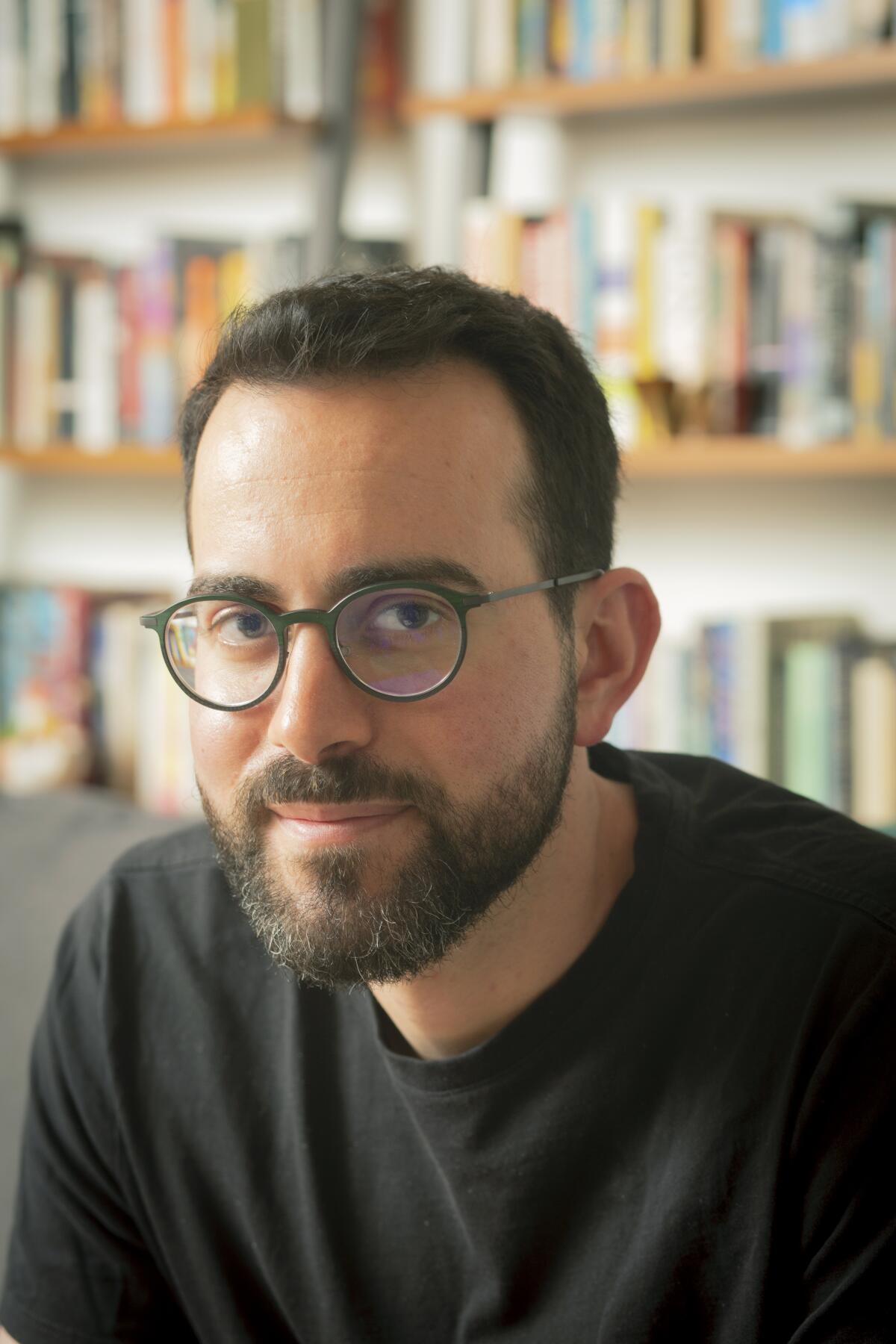 A bearded man wearing a dark crewneck shirt and glasses sits before shelves filled with books.