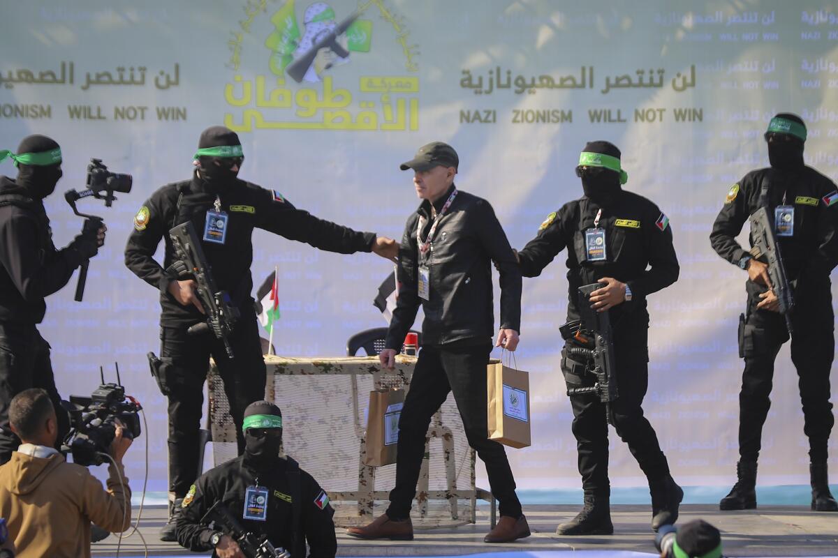 A black-clad man is escorted by black-clad Hamas fighters with faces mostly covered across a stage.