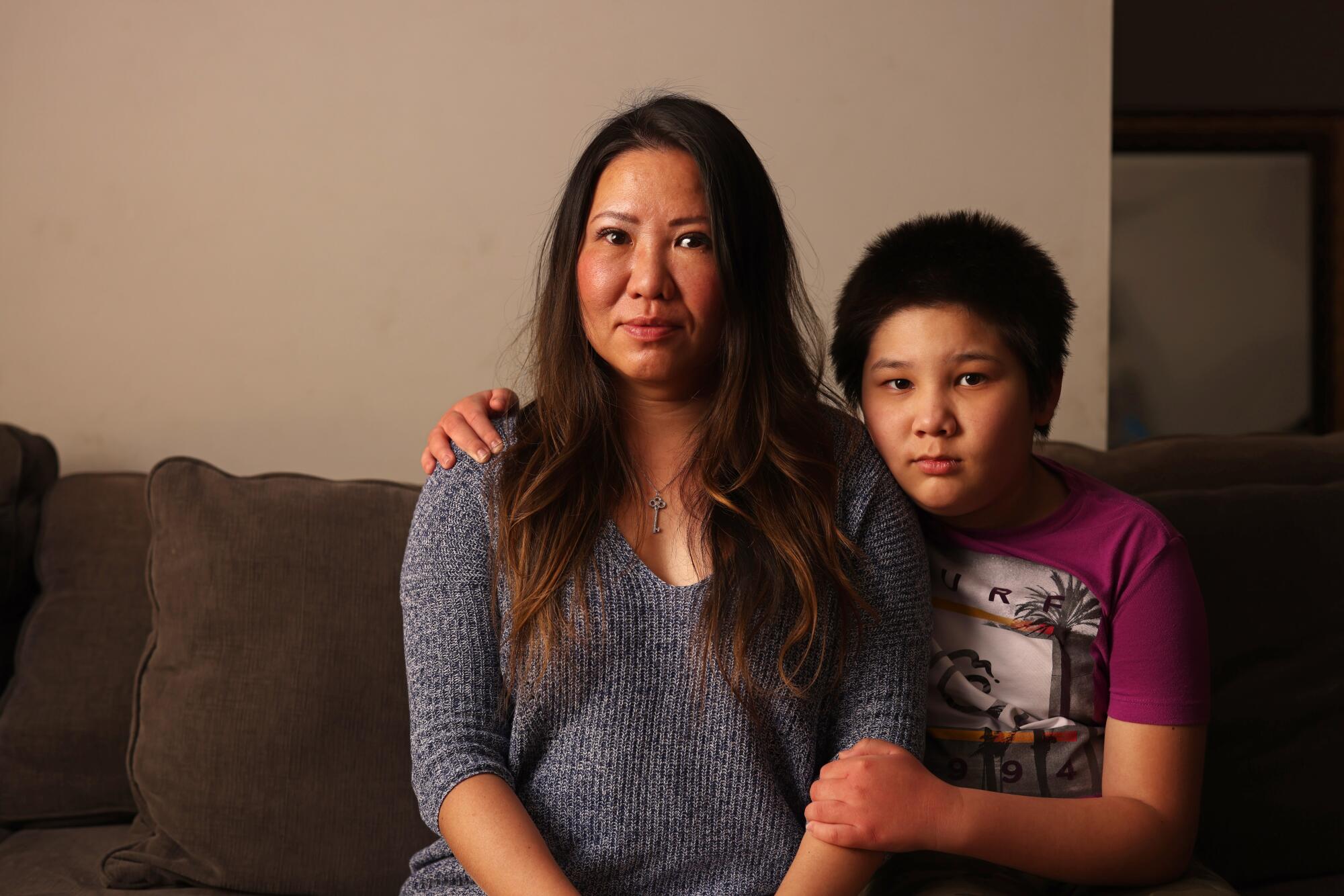 A woman sits on her couch, and her son sits next to her with his arms around her