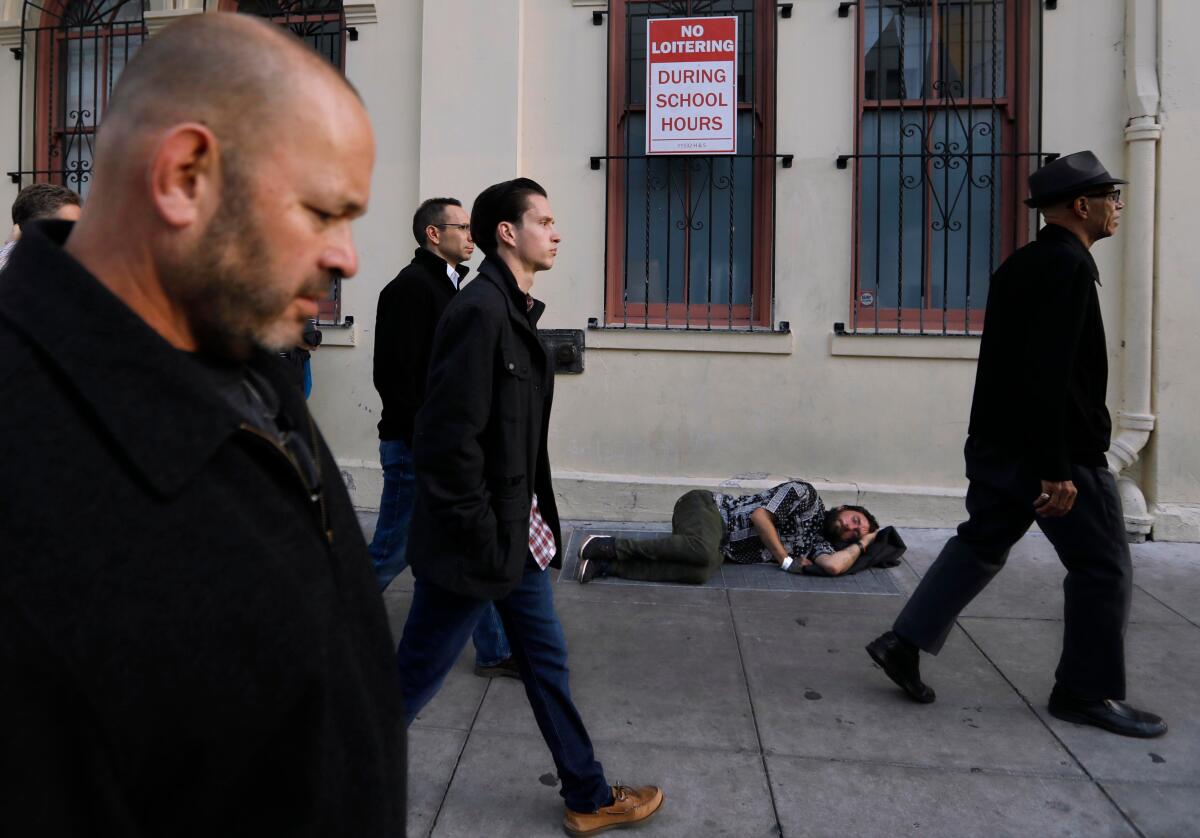 A man sleeps on a sidewalk in San Francisco as a group of four men, wearing black, walk past him.