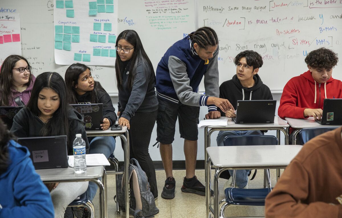 Standing between classroom desks, two peer mentors help other students during a class. 