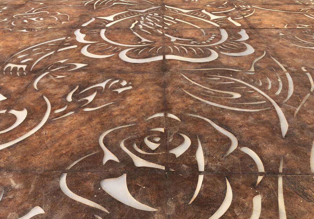 A detail view of corten steel plate lying on the ground show outlines of roses and a pair of praying hands.