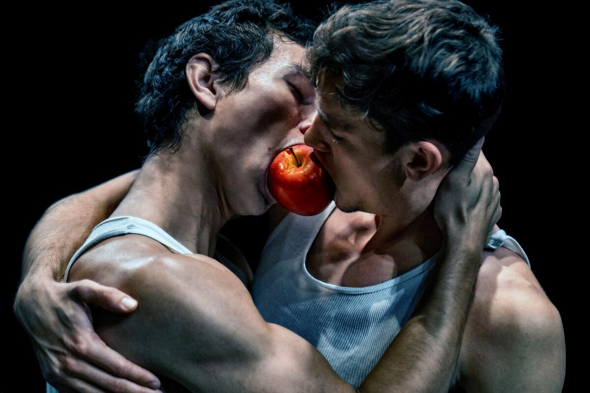 Two men embrace, biting into the same apple as if they are kissing.