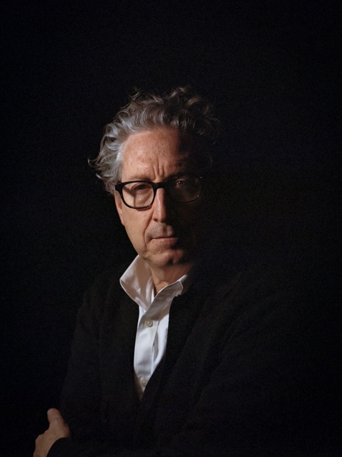 A portrait of a man with gray wavy hair and glasses in a white shirt and dark jacket.