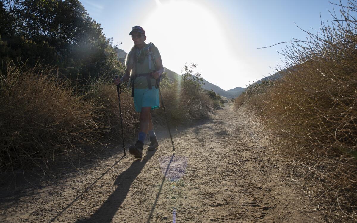 A person is seen from trail level hiking with trekking poles down a dusty, chapparal-lined trail, as the sun rises behind.