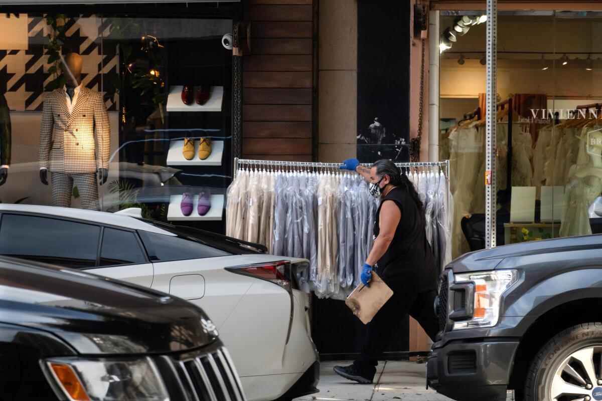 A person pushes a garment rack along a sidewalk near clothing stores with suits on display