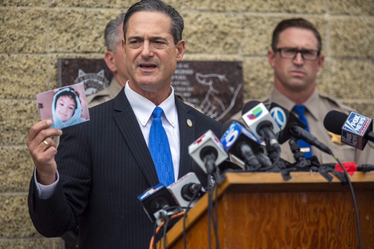 A man at a lectern covered with media microphones speaks and holds up a picture of a child.