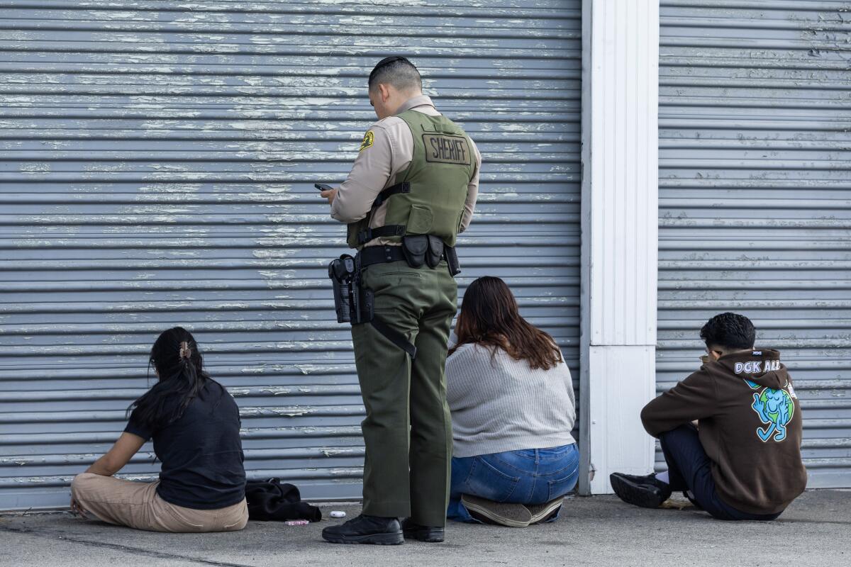 A uniformed sheriff's deputy in a ballistic vest stands beside three people seated facing corrugated metal garage gates