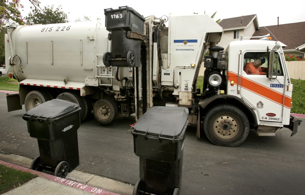 A garbage truck driver on a residential street looks up at a trash bin being lifted to be emptied into his truck