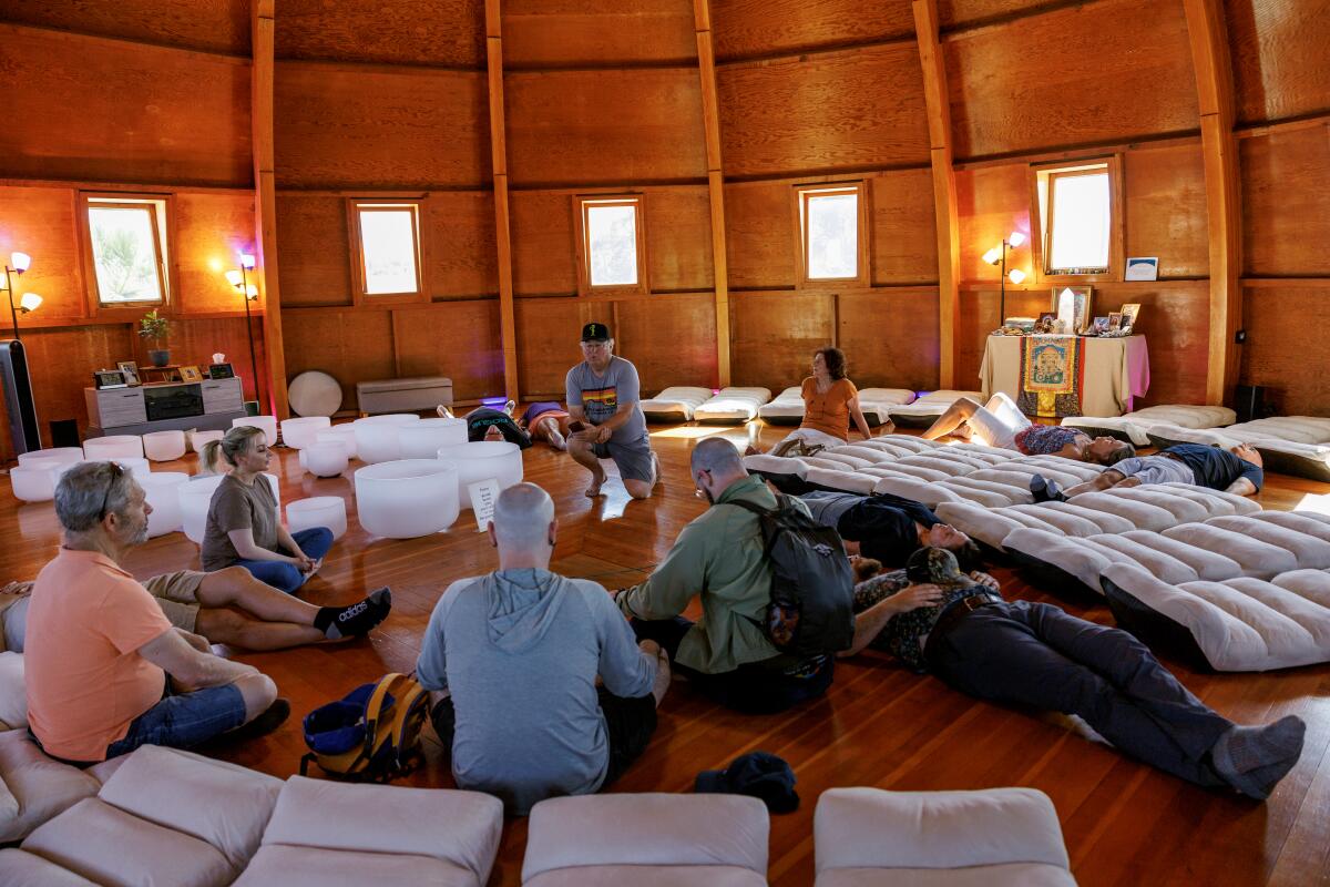 People inside a circular wooden building sit and lie on the floor. A man in the center plays tones for the visitors.