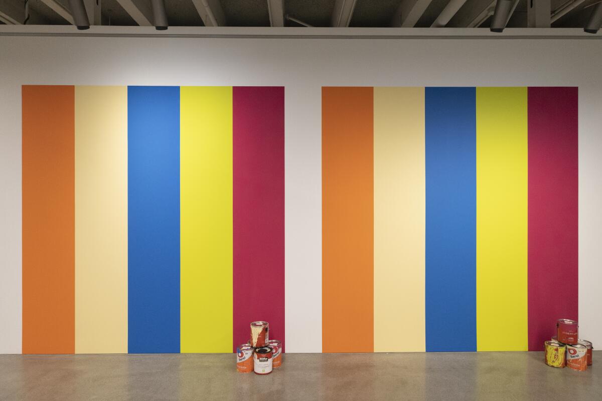 Two sets of five wide stripes of different colors — orange, cream, blue, yellow and red — are repeated on a gallery wall.