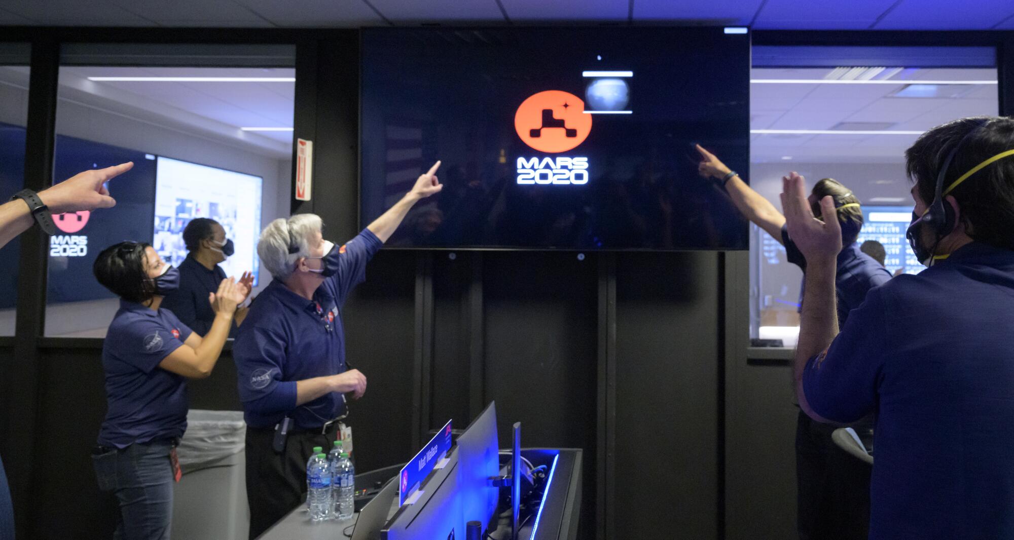Members of NASA's Perseverance rover team clap and point to a screen with a Mars 2020 logo
