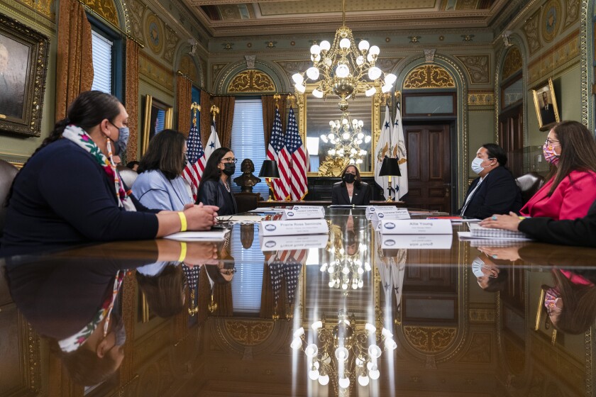 At a gleaming table beneath chandeliers, people in masks sit for a meeting.