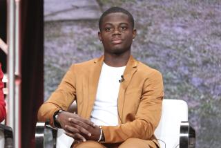 A Black man in a tan suit and a white t-shirt sitting in a white chair with his hands on the armrest