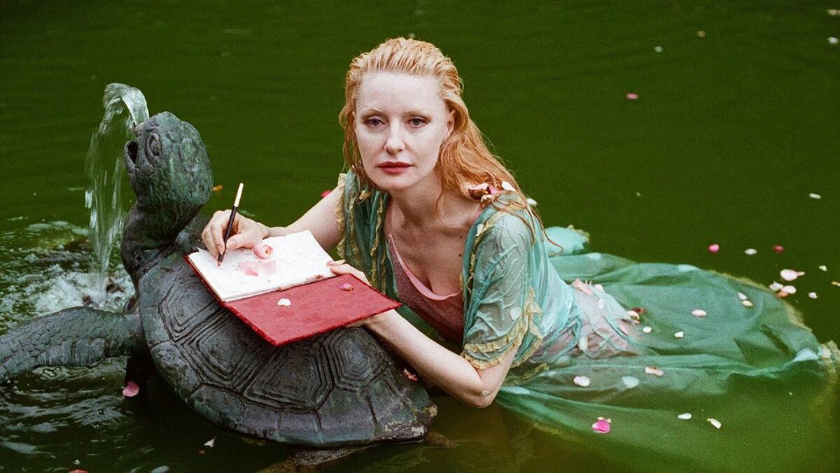 A woman in a green dress floats in a pond with a turtle sculpture and a journal.