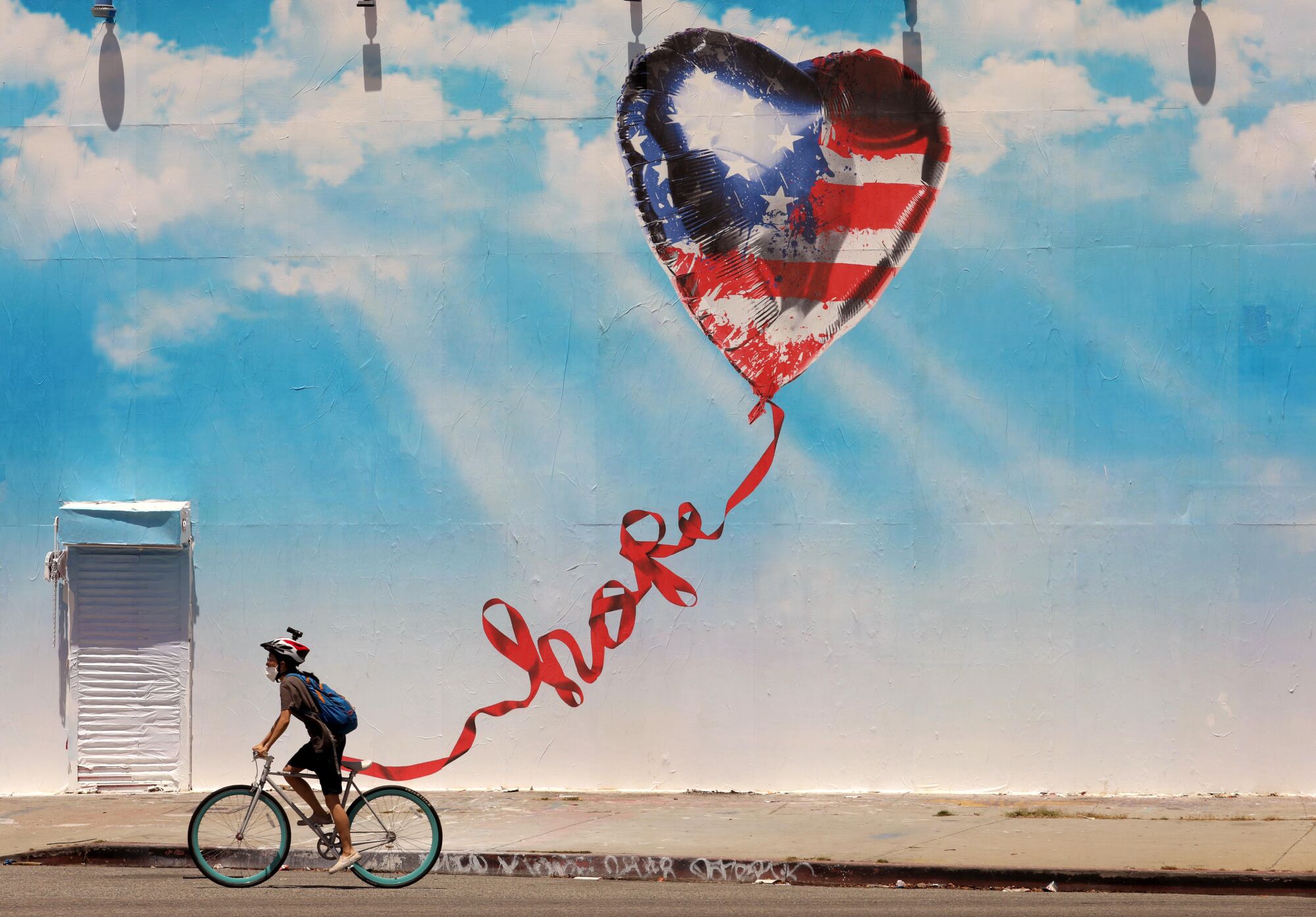 A cyclist rides past a mural of a heart-shaped U.S. flag balloon with a long ribbon that curls to spell out "hope."