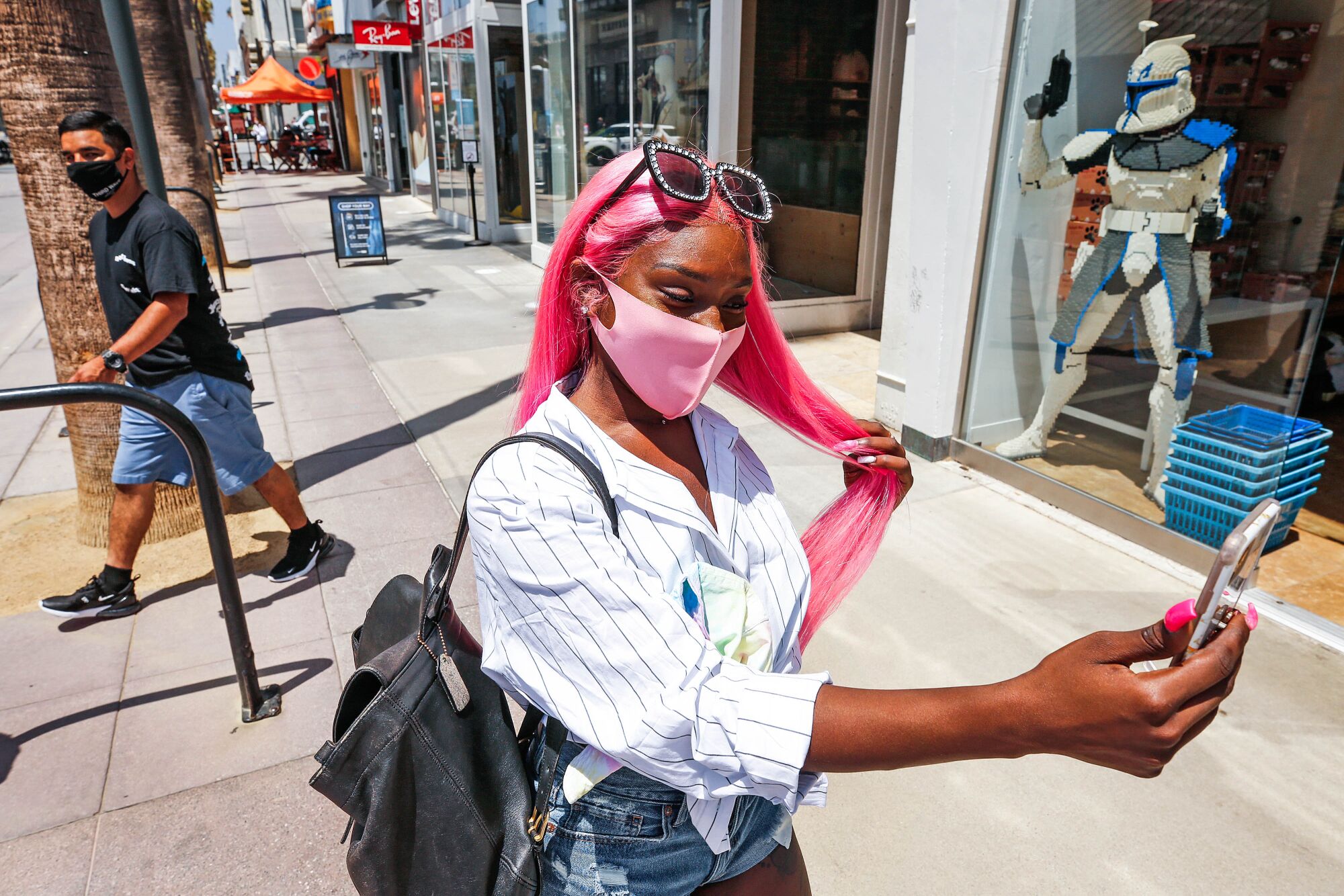 A woman with long pink hair and wearing a mask stands outside shops holding a phone.
