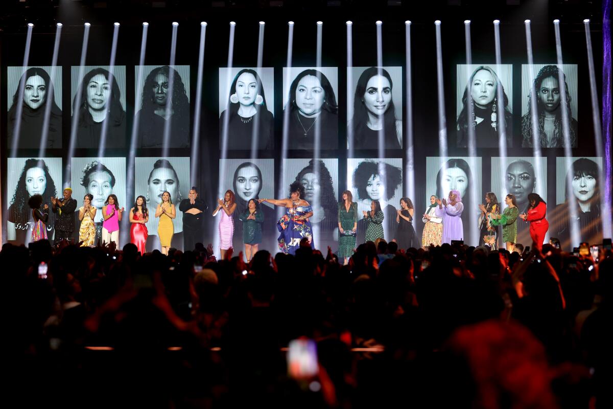 18 women on stager at an awards show with their photos projected behind them