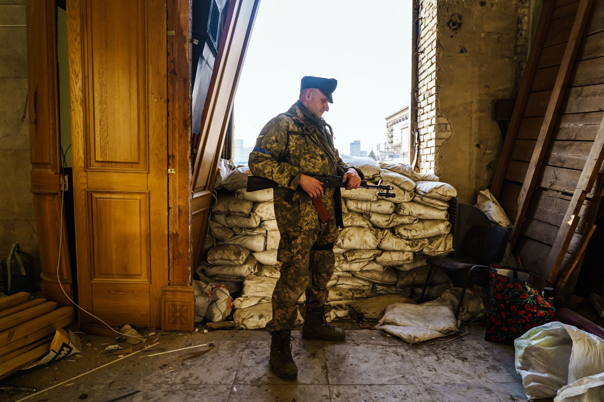 A male soldier in army fatigues armed with a rifle stands in a room with sandbags behind him in an entryway.