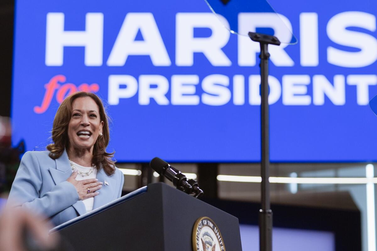 Kamala Harris places a hand on her chest as she speaks at a rally with "Harris for President" on a sign behind her