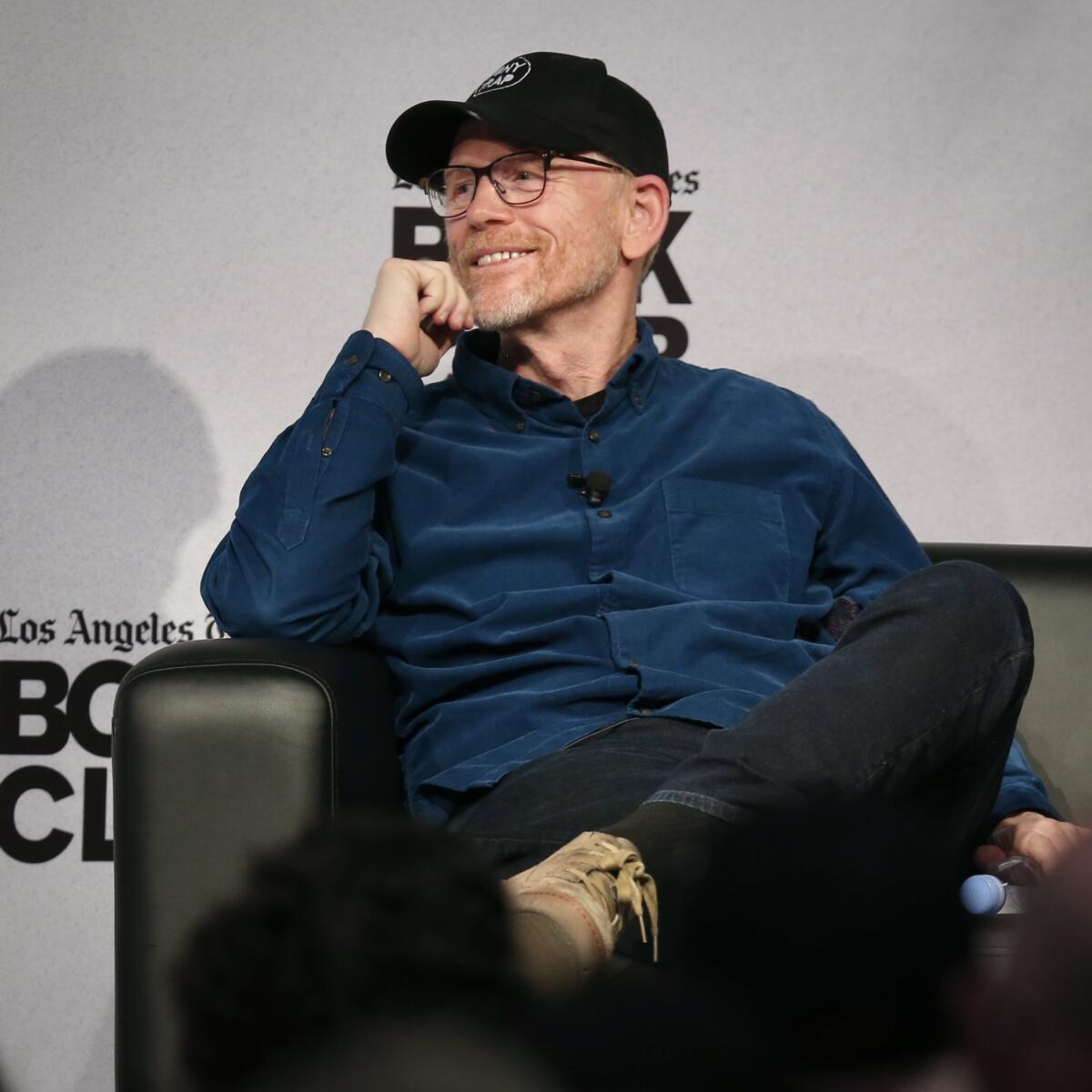 Ron Howard, seated and wearing ball cap and glasses, smiles.