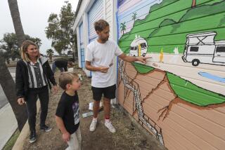 Swedish artist Jonas Claesson shows Wilson MacDowell, 6, son of Tate MacDowell, 39, a Cardiff resident who died of cancer in October, a camp fire scene that is part of a mural-painting inspired from a watercolor done by his father, titled "More Green Flashes", as Wilson's mother Lora Bodmer-MacDowell stands nearby the mural on the back wall of the Cardiff 7-Eleven convenience store on Tuesday, December 3, 2019 in Encinitas, California.