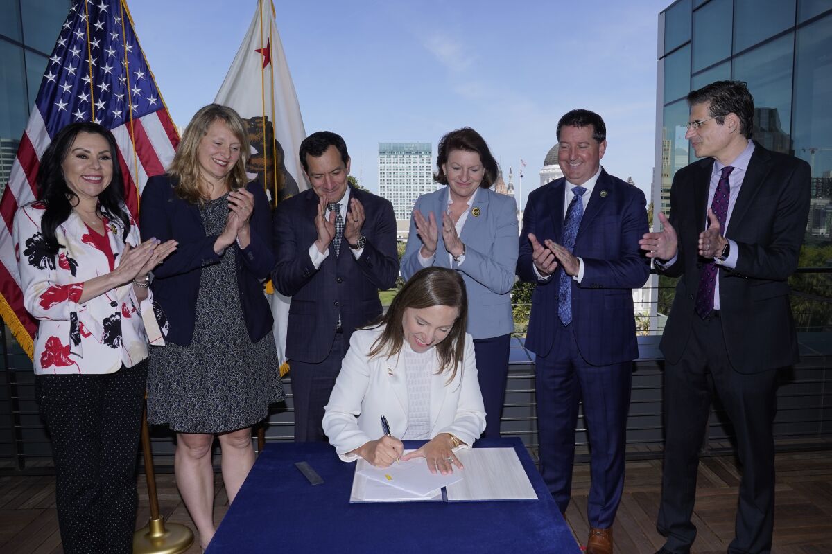 California Lt. Gov. Eleni Kounalakis signs a law as legislators applaud her from behind.