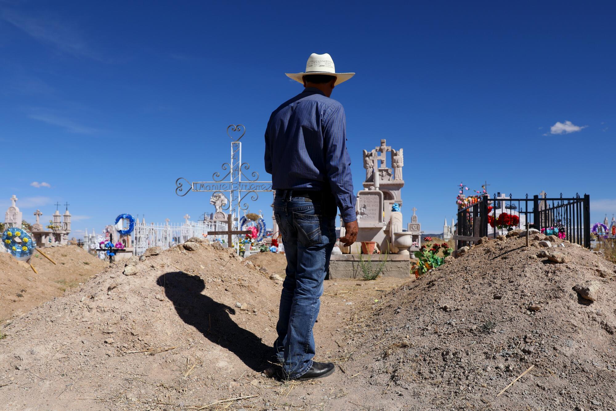 A man in a cowboy hat stands between two mounded graves in a dusty cemetery.