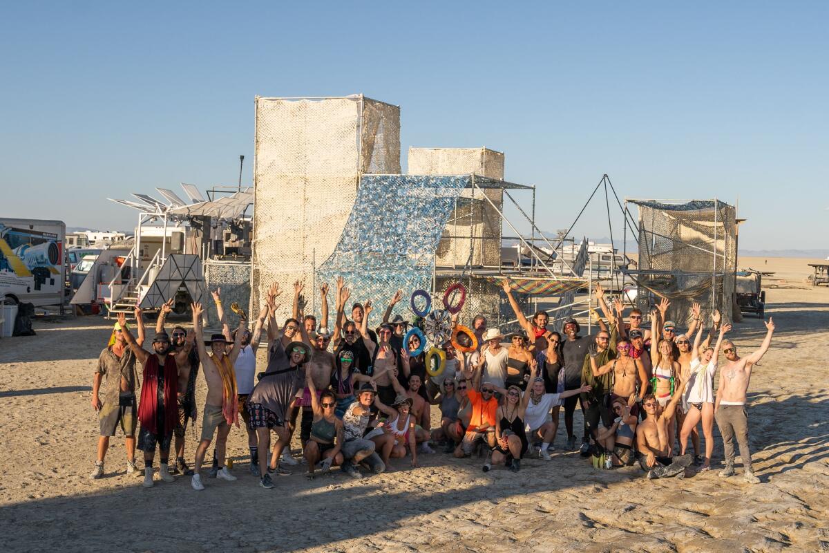 Une photo de groupe de plusieurs personnes saluant sur le sable avec des structures derrière elles à Burning Man.