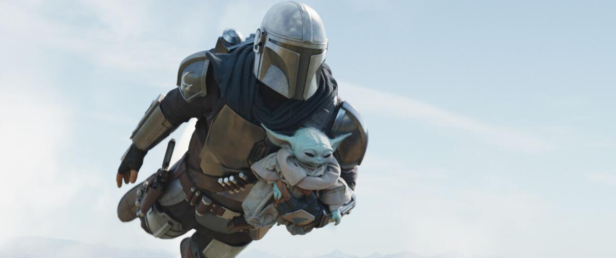 A man in a Mandalorian costume and helmet flying through the air while carrying Grogu, a.k.a. Baby Yoda