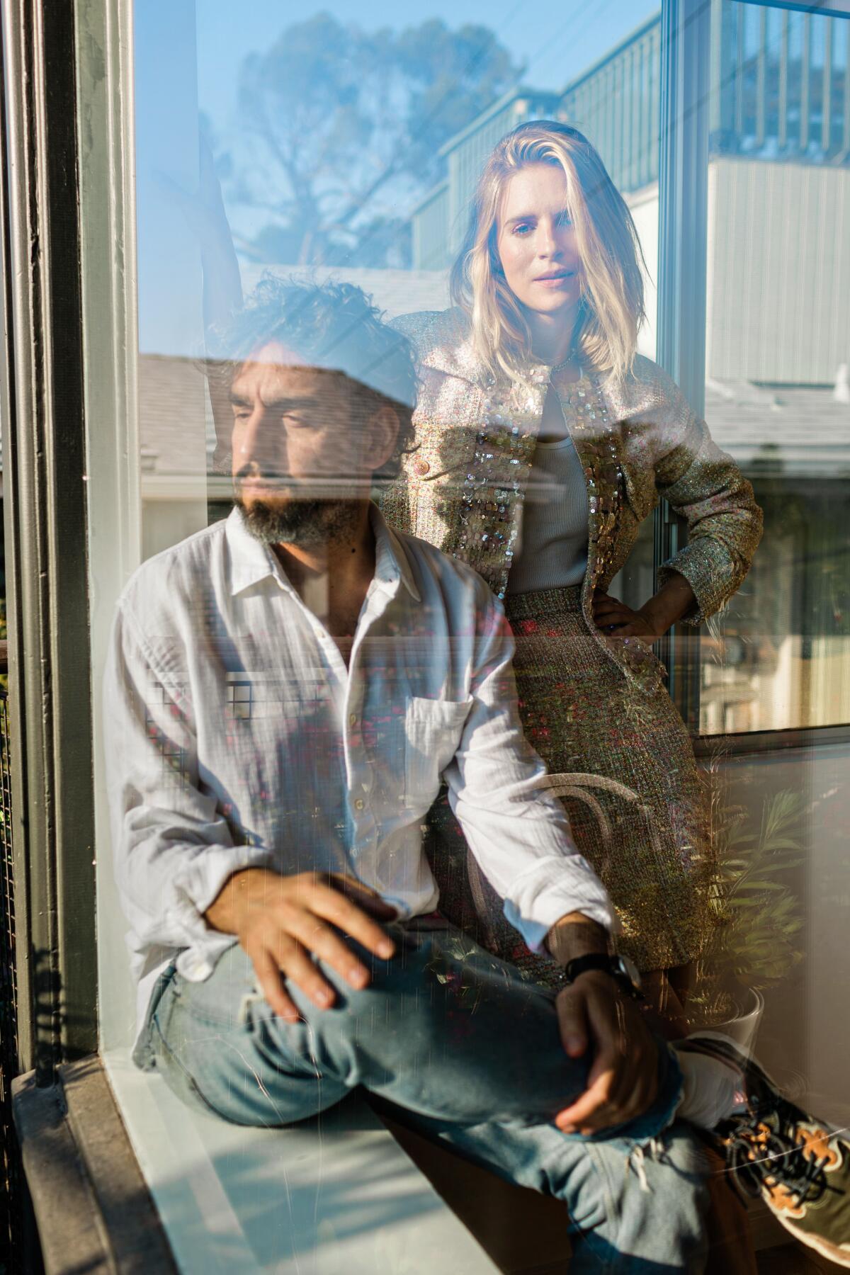 Seen through a picture window, a bearded man in a white shirt looks out as a blond woman behind him looks into the lens.