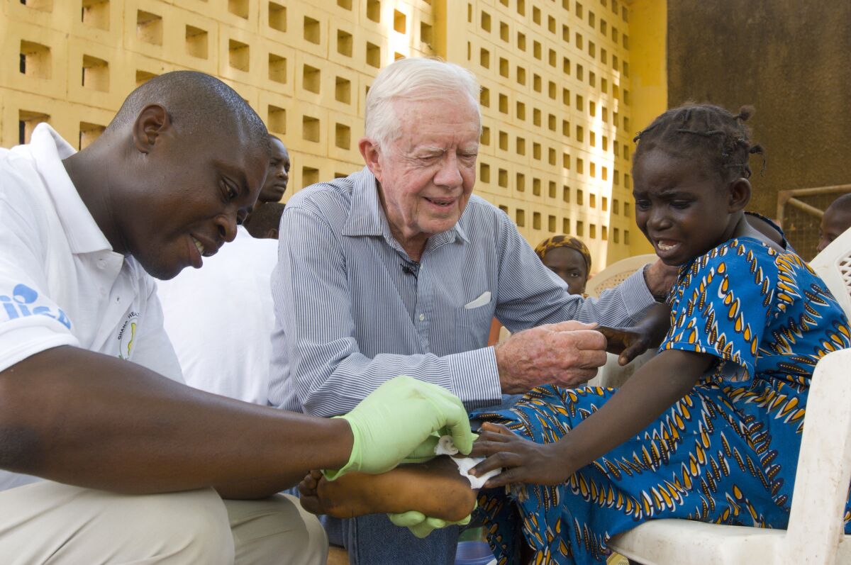Jimmy Carter, center, consoles a child grimacing as a health worker dresses a wound on her hand