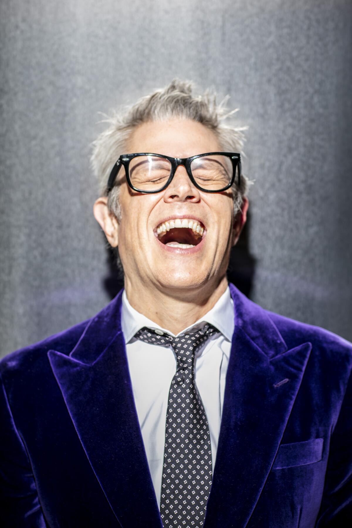 A man in a purple suit jacket laughs for a portrait.