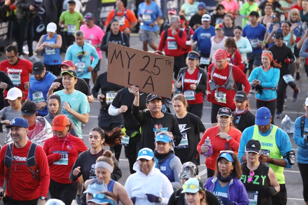 People in running gear crowd together as they run. One runner holds a sign that says "My 25th LA."