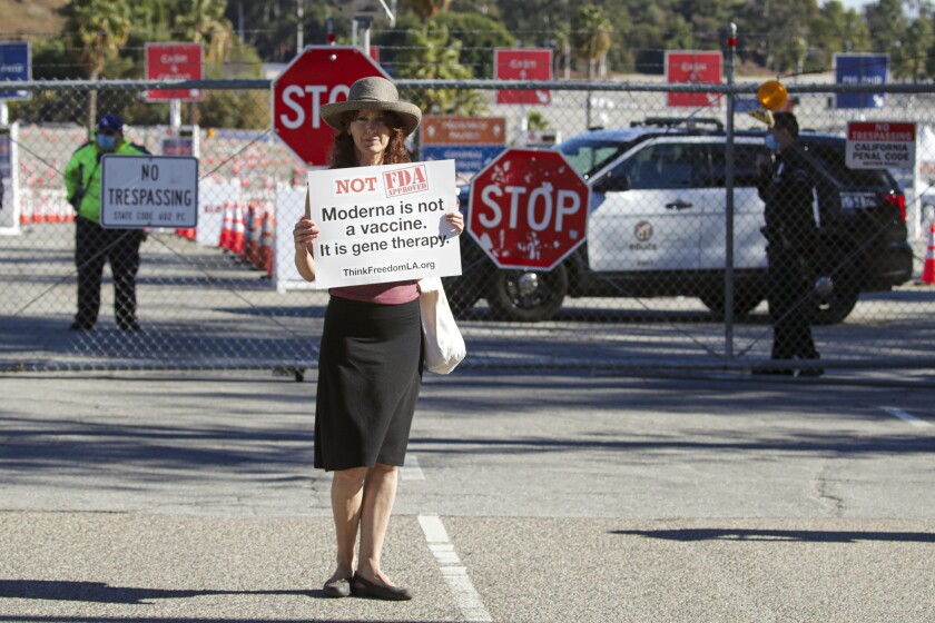 A woman stands near a chain link fence with a sign that says "Modern is not a vaccine. It is gene therapy."