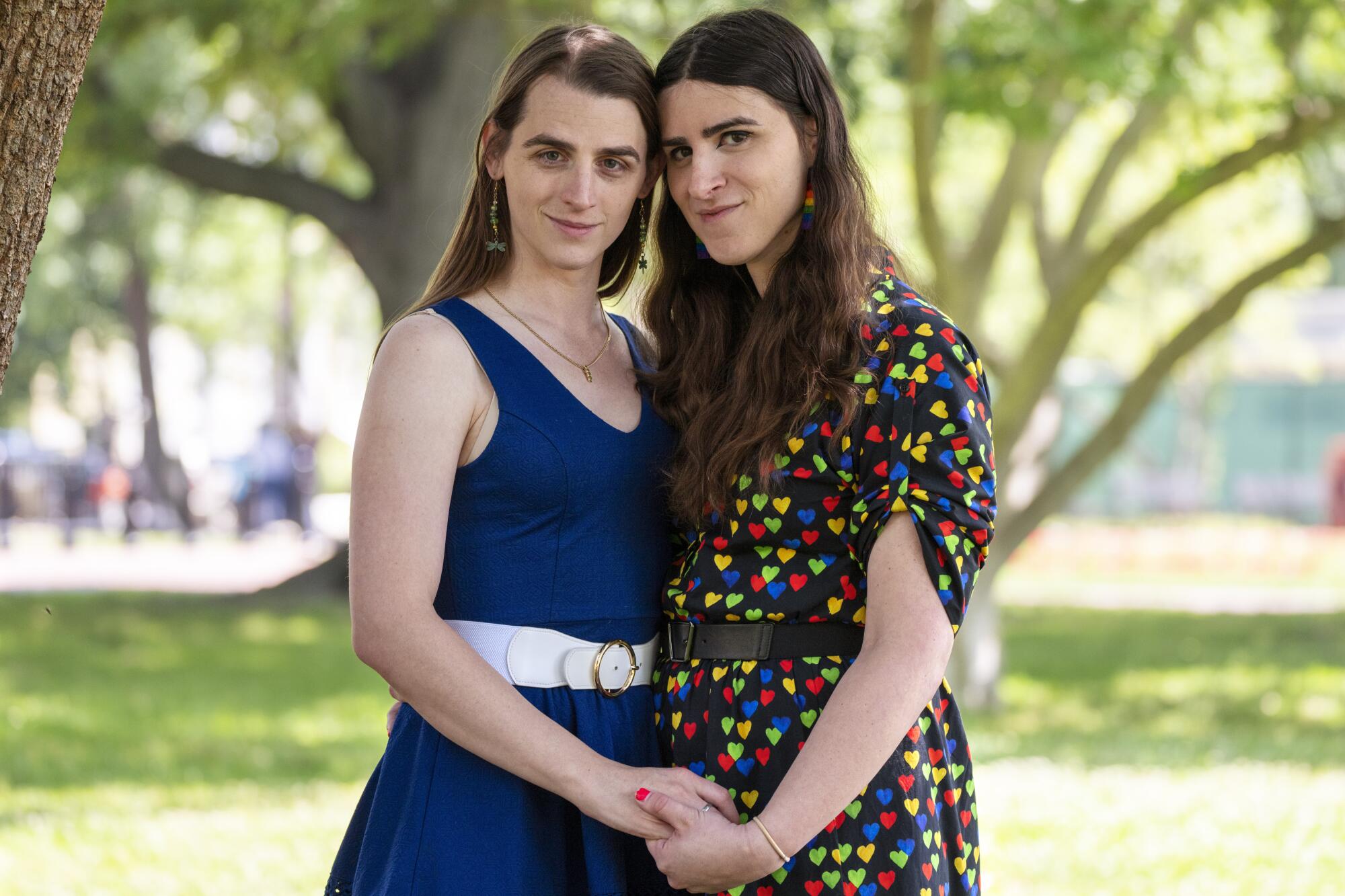 Zooey Zephyr and Erin Reed lean in and hold hands for a photo in a park-like setting with large trees.