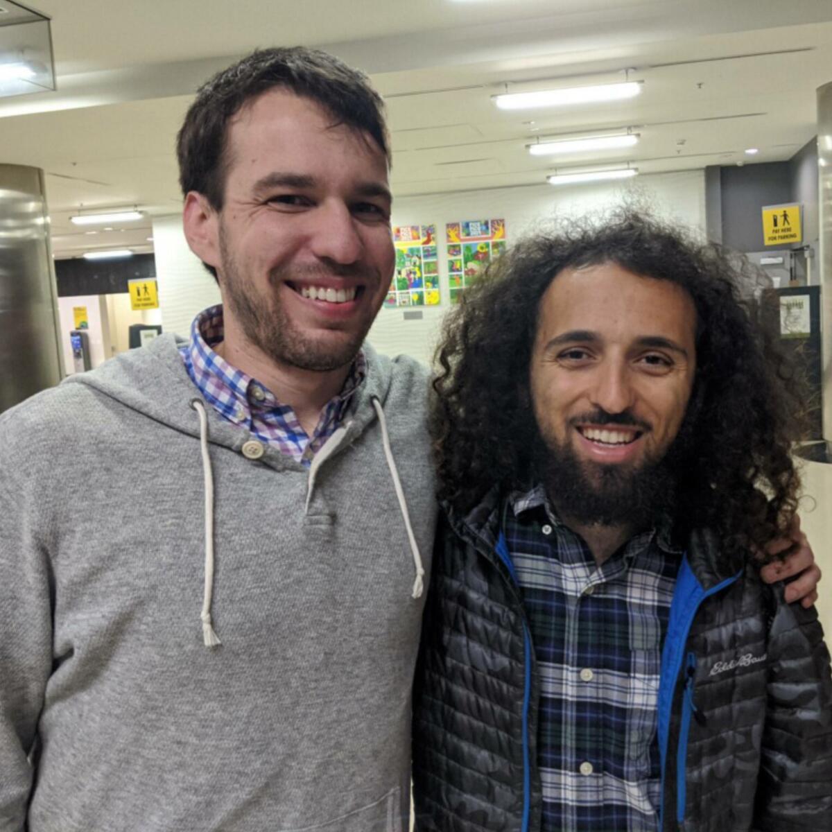 Two friends smile for the camera, one with his arm around the other's shoulder