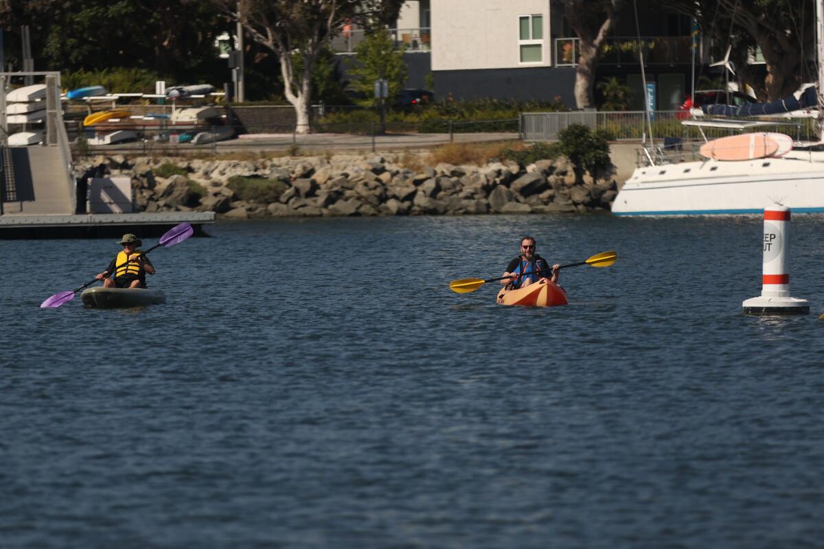 Two people paddling solo near a buoy, a large boat and the shoreline not far behind them