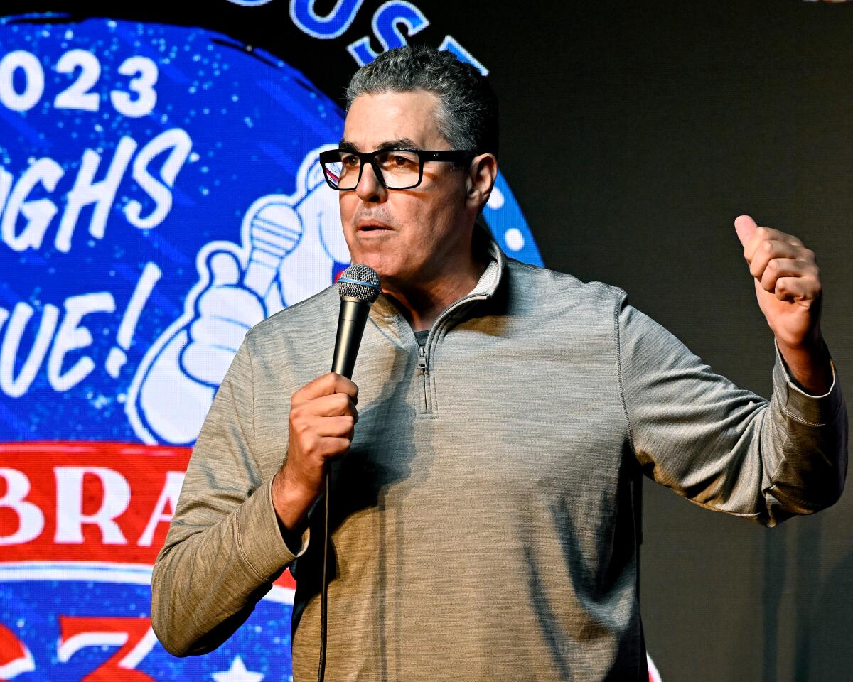 Adam Carolla speaking into a microphone and lifting one hand while clad in a casual long-sleeve gray shirt