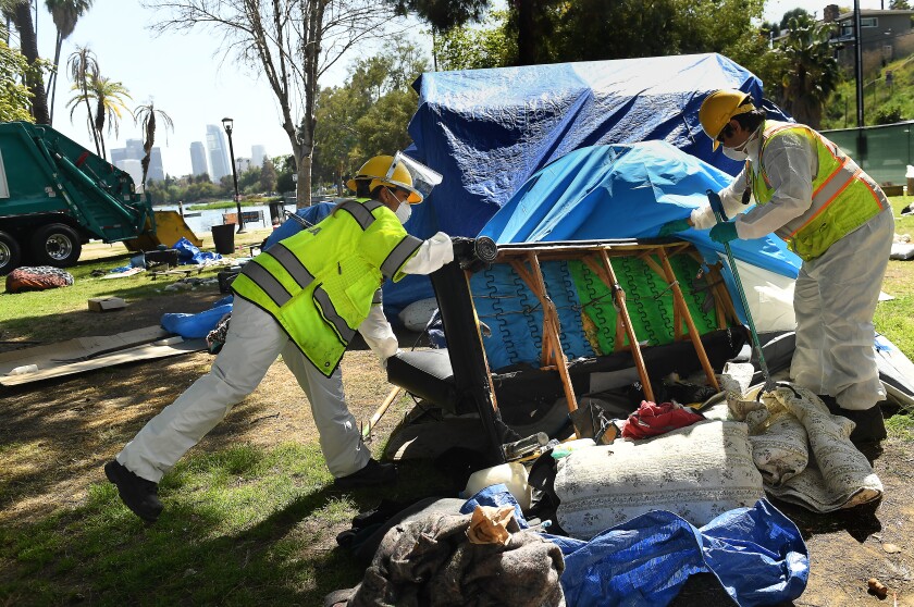Two workers in high-visibility vests lift a piece of furniture in a park littered with tarps, blankets and other objects