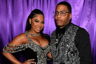 Ashanti smiling in a glittering gown and standing with Nelly who has on a matching glittery vest, black shirt and glasses