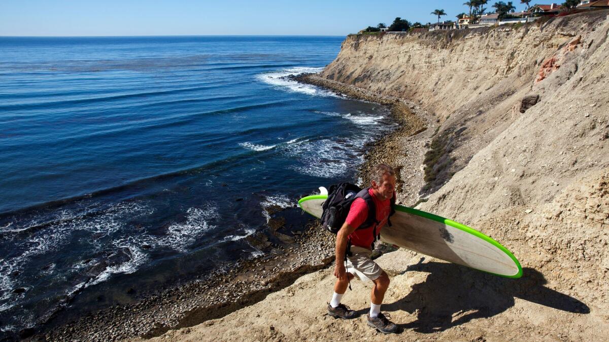 A man who declined to give his name carries a surfboard up a trail as city contractors demolish a patio built by local surfers.