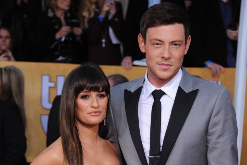 Lea Michele is wearing a pink strapless dress and is posing with Cory Monteith who is in a gray suit with black trim
