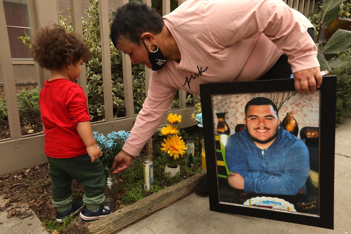 A small child watches as his grandmother holds a picture of her son next to an outdoor memorial.