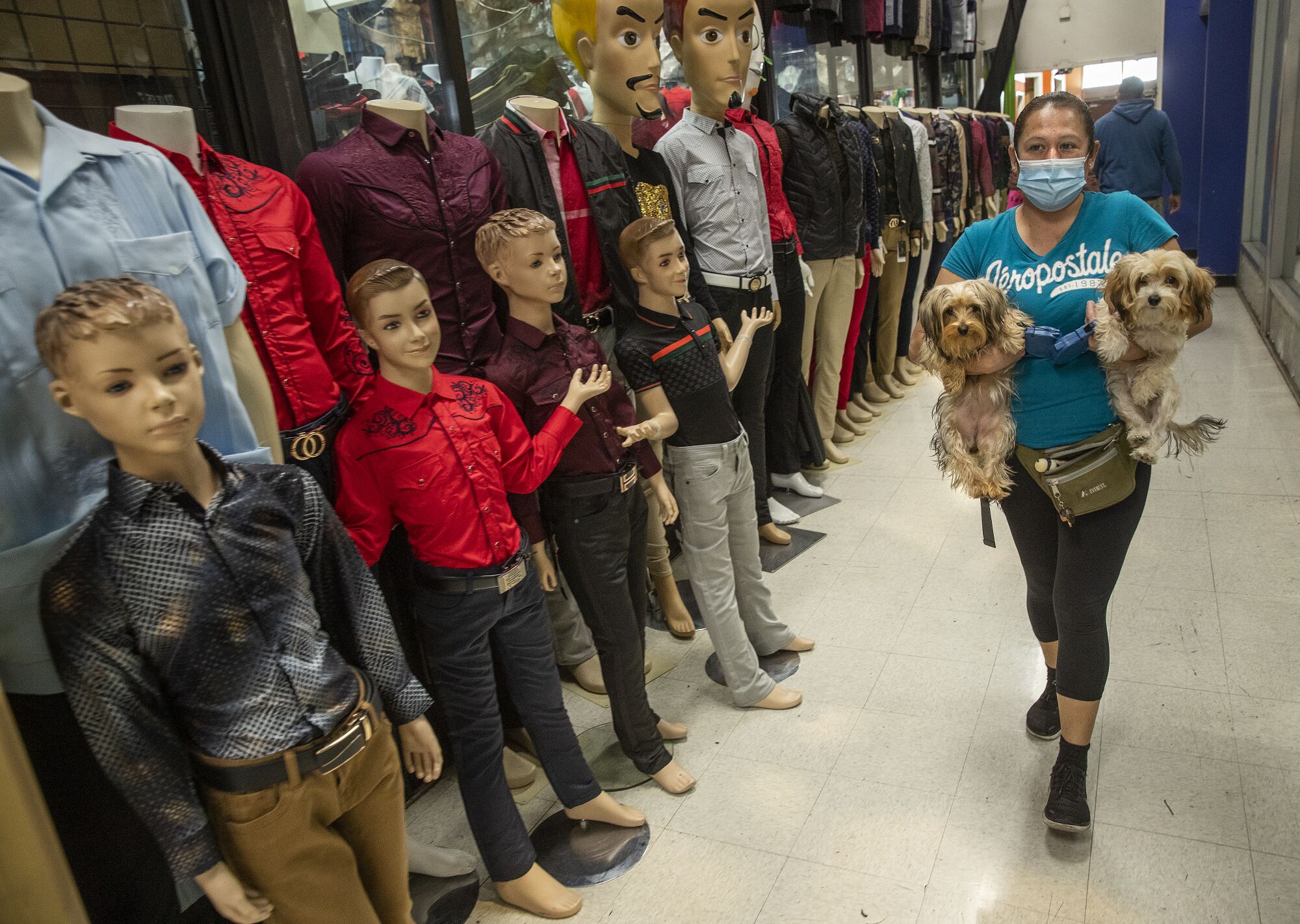 A woman, carrying a dog under each arm, walks down a corridor past mannequins of boys. 