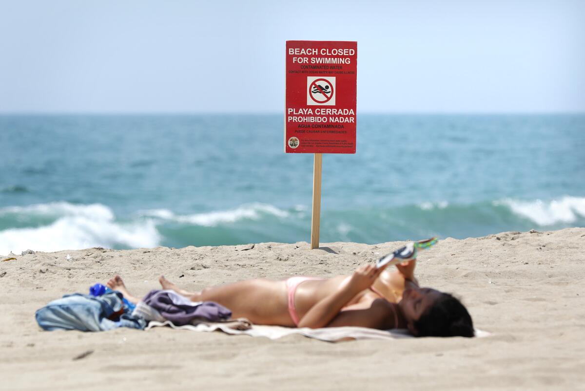 A beachgoer sunbathes near a sign warning people to stay out of the water at Dockweiler State Beach.